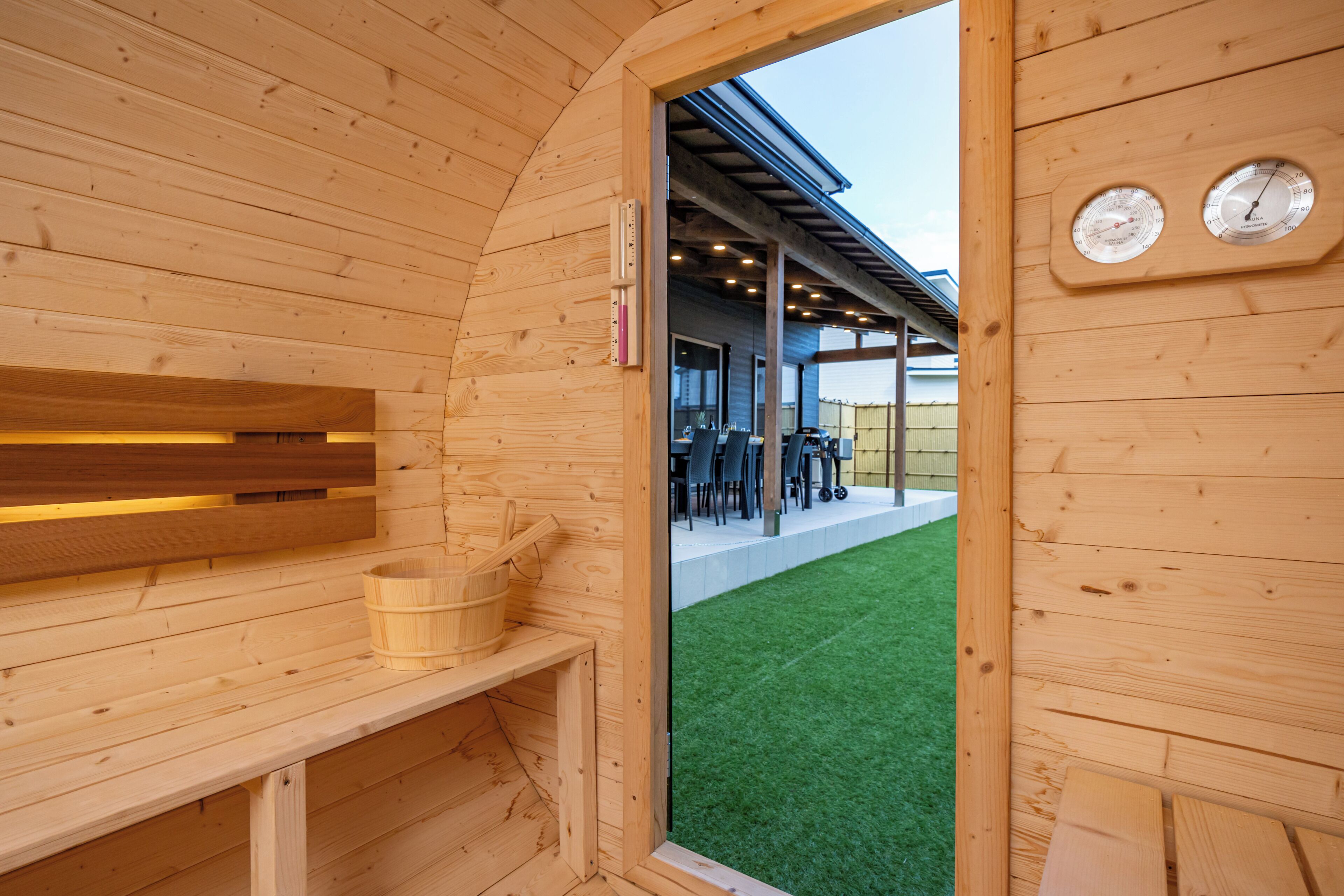 A wooden sauna with a bench, a wooden bucket, and a door leading to an outdoor area with a covered seating area and a green lawn.