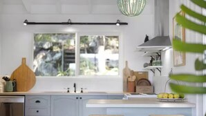 Une cuisine avec une fenêtre, des meubles blancs et un luminaire suspendu, ainsi que deux chaises en osier.