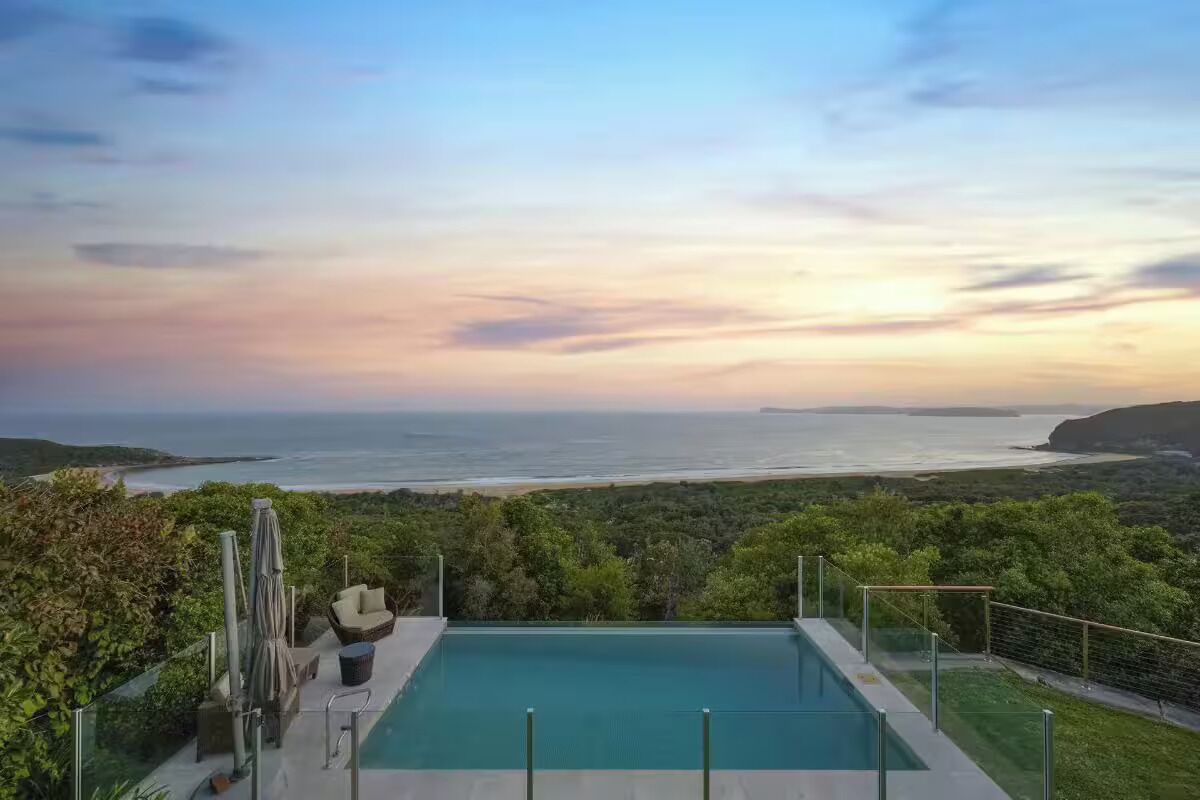 Une piscine rectangulaire à débordement, dotée d’une balustrade en verre, entourée de verdure et offrant une vue sur un paysage côtier.