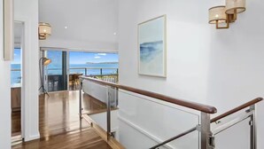 Un interior amplio con piso de madera, una escalera con barandilla de vidrio y vista al mar a través de grandes ventanales.