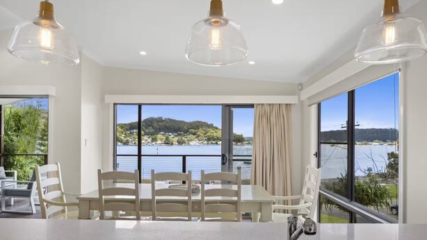 A modern kitchen with a large window overlooking a lake, a dining area with chairs, and pendant lights. - Luxury Villas Araluen Waterfront Getaway (Walk to the beach) (Central Coast)