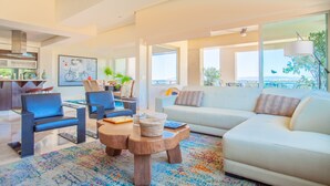 Ein modernes Wohnzimmer mit einem weißen Sofa, blauen Sesseln und einem Couchtisch. Im Hintergrund ist eine Küche mit einem großen Fenster zu sehen.