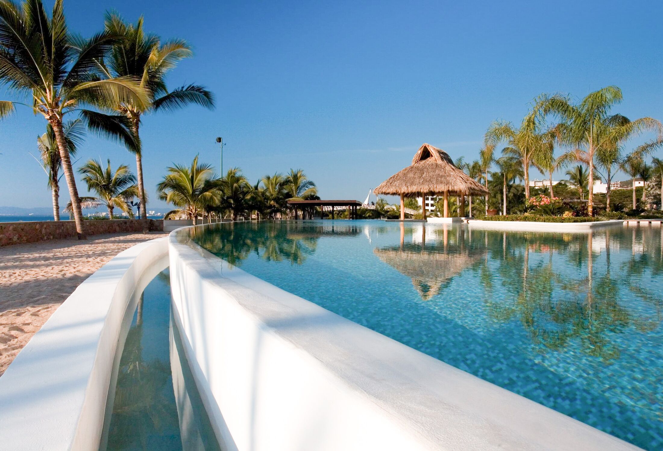 A clear swimming pool with a thatched-roof structure in the background, surrounded by palm trees and a beach.