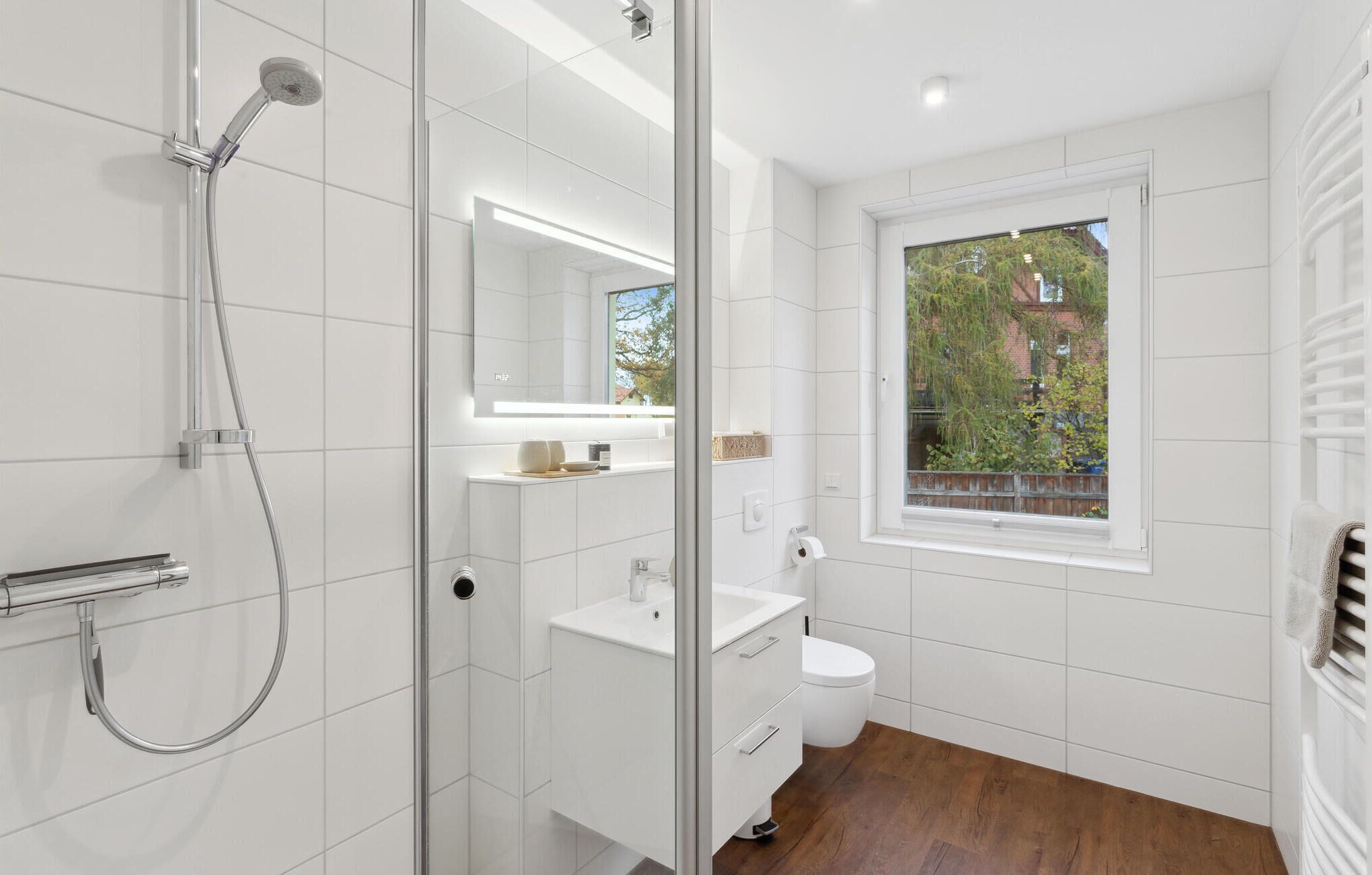 Ein modernes Badezimmer mit Duschkabine aus Glas, einem weißen Waschbecken und einer Toilette. Ein Fenster bietet Blick ins Freie.