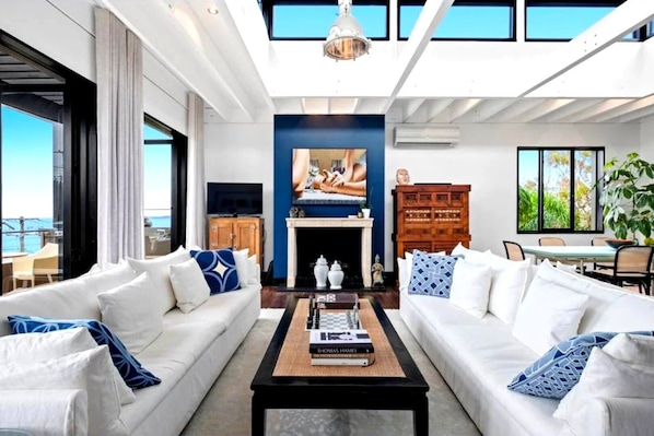 A modern living room with two white sofas, a coffee table with books, a fireplace, and a large window overlooking the ocean.