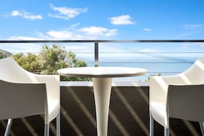 A balcony with a white table and two white chairs, overlooking a scenic view of the sea and a bridge.