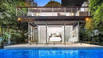 Enjoy a bedroom with a large bed, a covered outdoor seating area, and a spacious outdoor swimming pool surrounded by lush greenery.