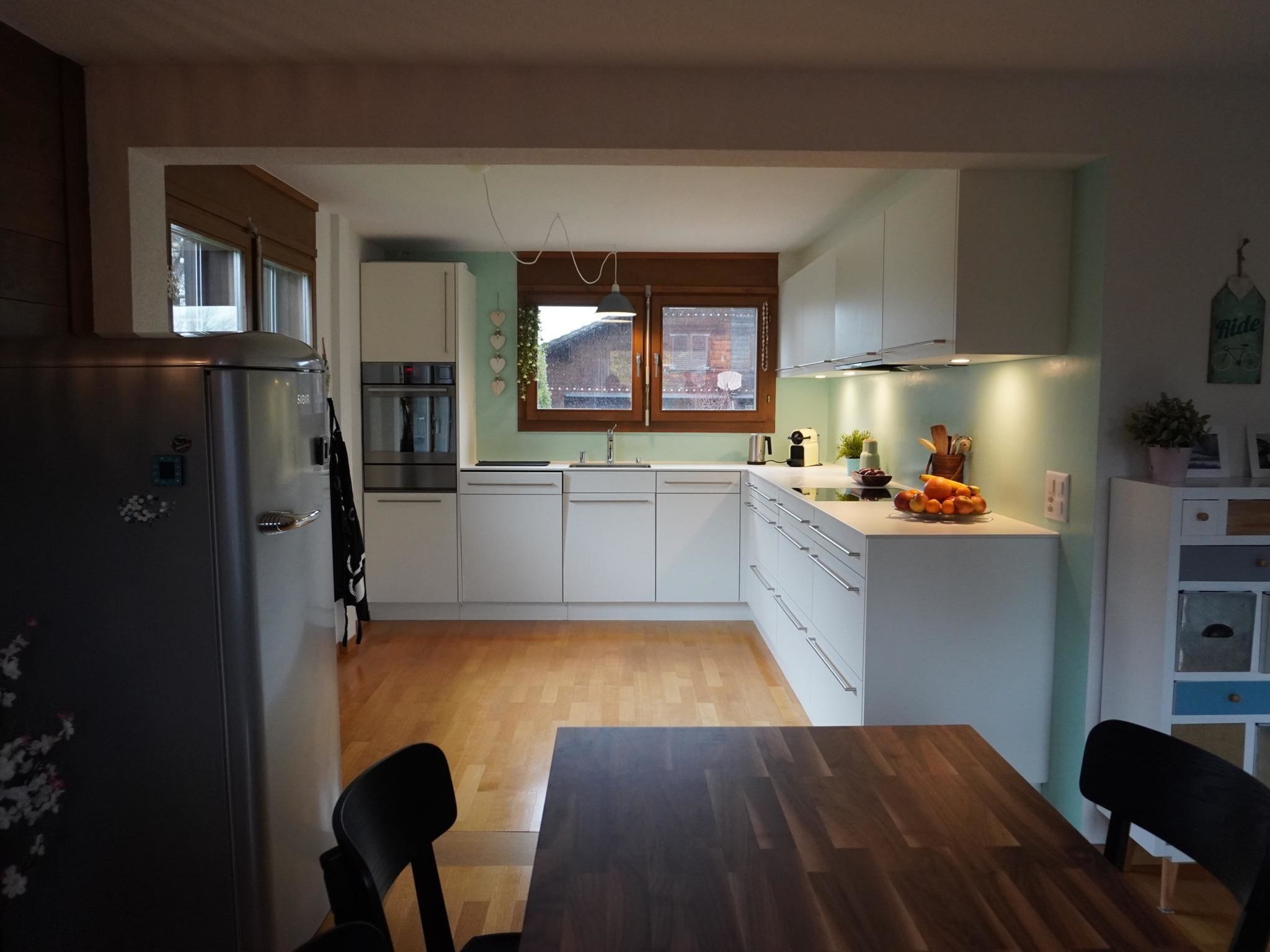 Moderni keittiö, jossa on valkoiset kaapit, puinen ruokapöytä ja jääkaappi.