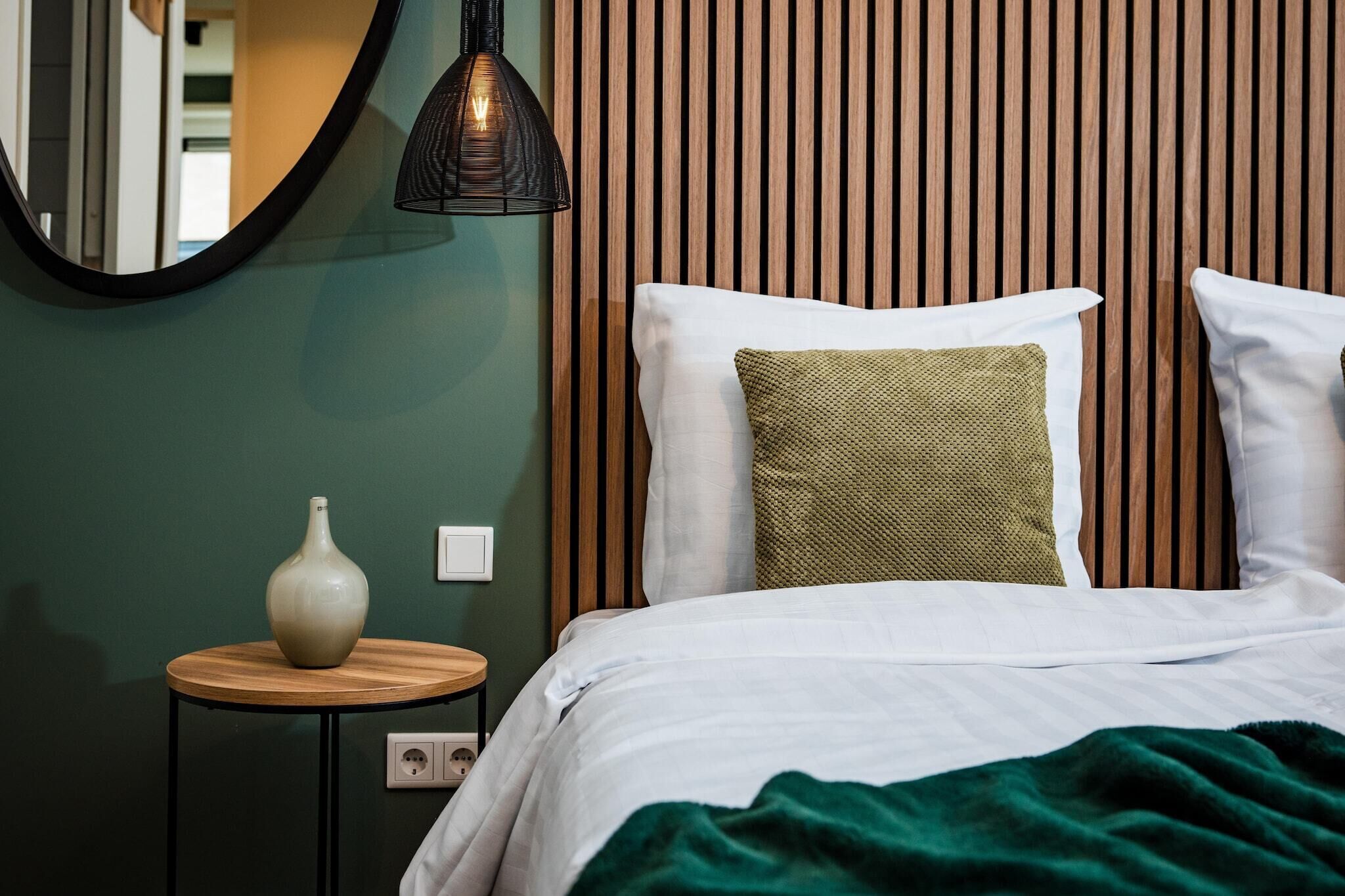Un letto rifatto con cura, con biancheria bianca e due cuscini, uno verde e uno bianco, appoggiato a una parete con listelli di legno verticali. A sinistra, si vede un tavolo rotondo in legno con un vaso bianco e una lampada a sospensione nera.