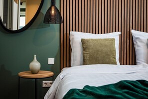 A neatly made bed with white bedding and two pillows, one green and one white, against a wall with vertical wooden slats. A round wooden table with a white vase and a black pendant light is visible to the left.