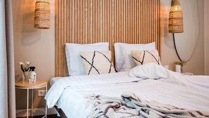 Een net opgemaakt bed met witte lakens en decoratieve kussens, een klein nachtkastje met een vaas en een spuitfles, en twee rieten hanglampen.