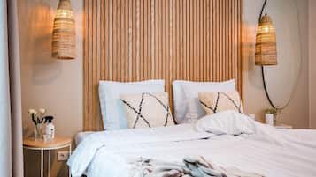 A neatly made bed with white linens and decorative pillows, a small bedside table with a vase and a spray bottle, and two wicker pendant lights.
