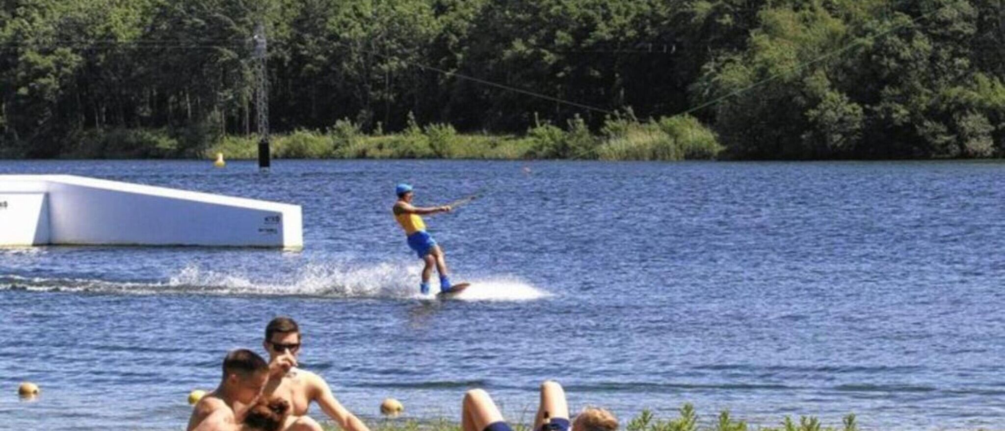Una persona practica wakeboard en un lago, con un barco al fondo.