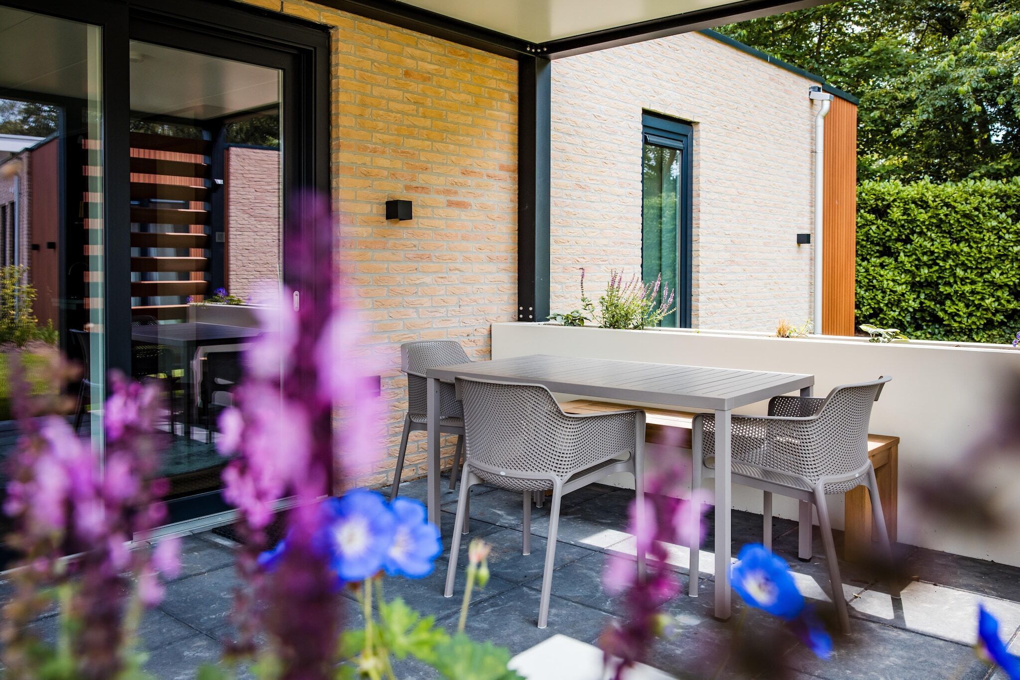 Een terras buiten met een tafel en stoelen, een glazen deur die leidt naar een trap, en een bakstenen muur.