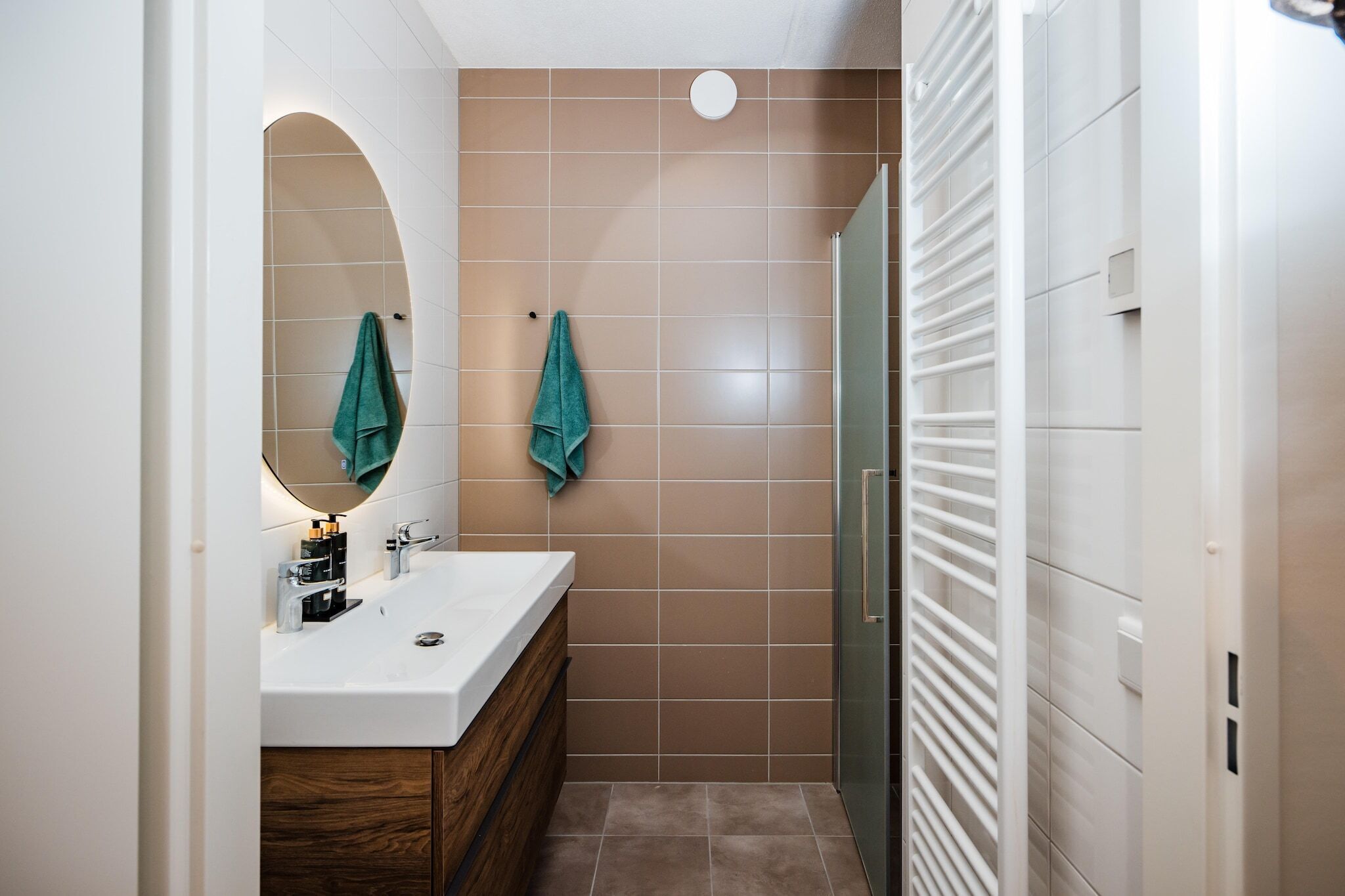 A modern bathroom with a white sink, wooden vanity, two green towels, and a glass shower enclosure.