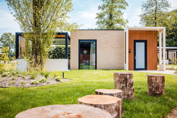 A modern building with a brick facade and a wooden door, surrounded by a well-maintained garden with grass and trees. - Chalet in Voorthuizen near Veluwe Nature (Voorthuizen)