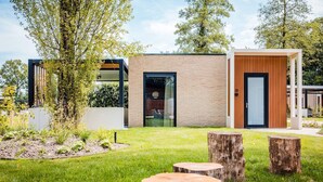 Een modern gebouw met een bakstenen gevel, een glazen deur en een houten deur. Op de voorgrond staan bomen en een gazon met houten blokken.
