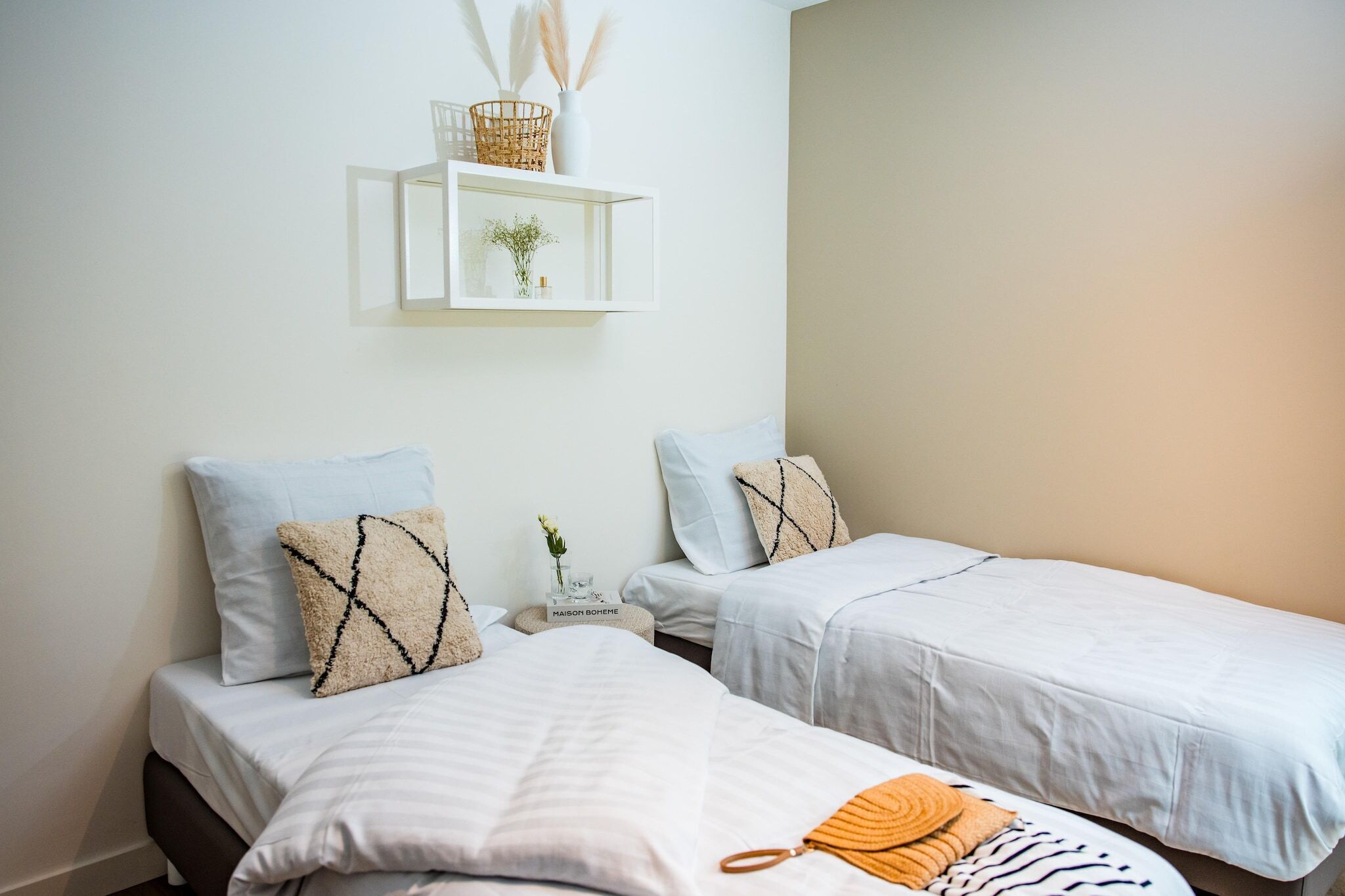 Habitación de hotel con dos camas individuales, una mesita de noche con un florero y una vela, y un estante en la pared con objetos decorativos.
