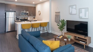Et moderne køkken med stålemmer, en central ø med barstole, et opholdsområde med sofa og TV-reol, samt en dekorativ plante.