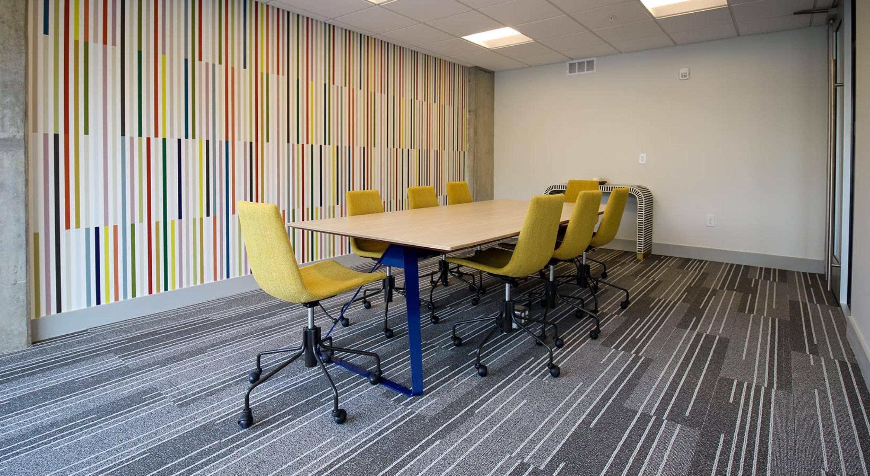 Une salle de conférence avec une longue table, des chaises jaunes et un mur rayé aux couleurs vives.