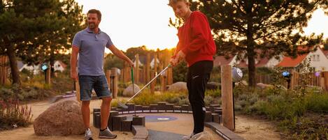 A person in a red sweater and black pants holding a golf club, standing on a sand-based putting green with a wooden border.