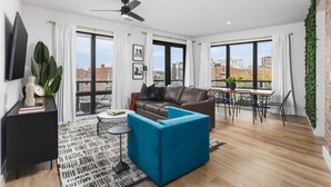 A modern living room with a leather sofa, a blue ottoman, a coffee table, and a TV on a black stand. There is a dining area with a table and chairs, and large windows offering a view of the city.