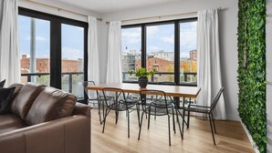 A modern living room with a brown leather sofa, a wooden dining table with chairs, large windows with white curtains, and a green ivy wall.