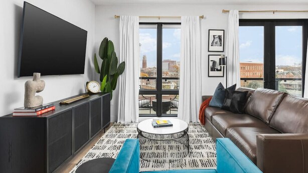 A modern living room with a leather sofa, a round coffee table, a flat-screen TV mounted on the wall, and a balcony with a city view.