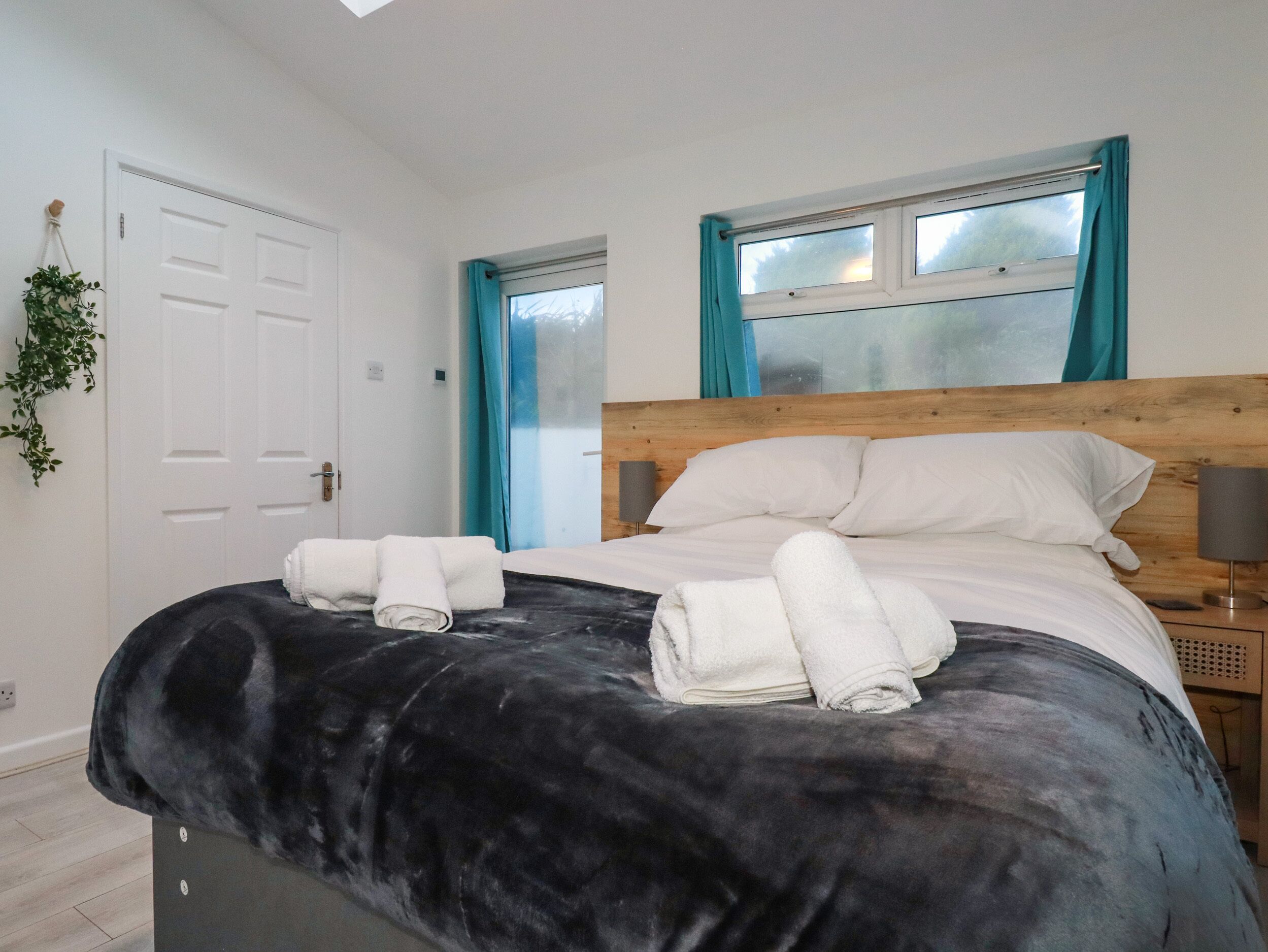 Een netjes opgemaakt bed met witte lakens en twee opgerolde handdoeken, een houten hoofdbord, een nachtkastje met een lamp en een raam met blauwe gordijnen.