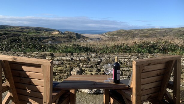 Une table en bois, accompagnée de deux chaises, est placée face à un paysage pittoresque composé de collines et d’une plage.