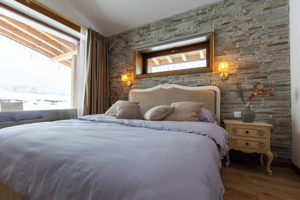 A bedroom with a stone wall, a wooden ceiling, a bed with a headboard, two bedside tables with lamps, and a window with curtains.