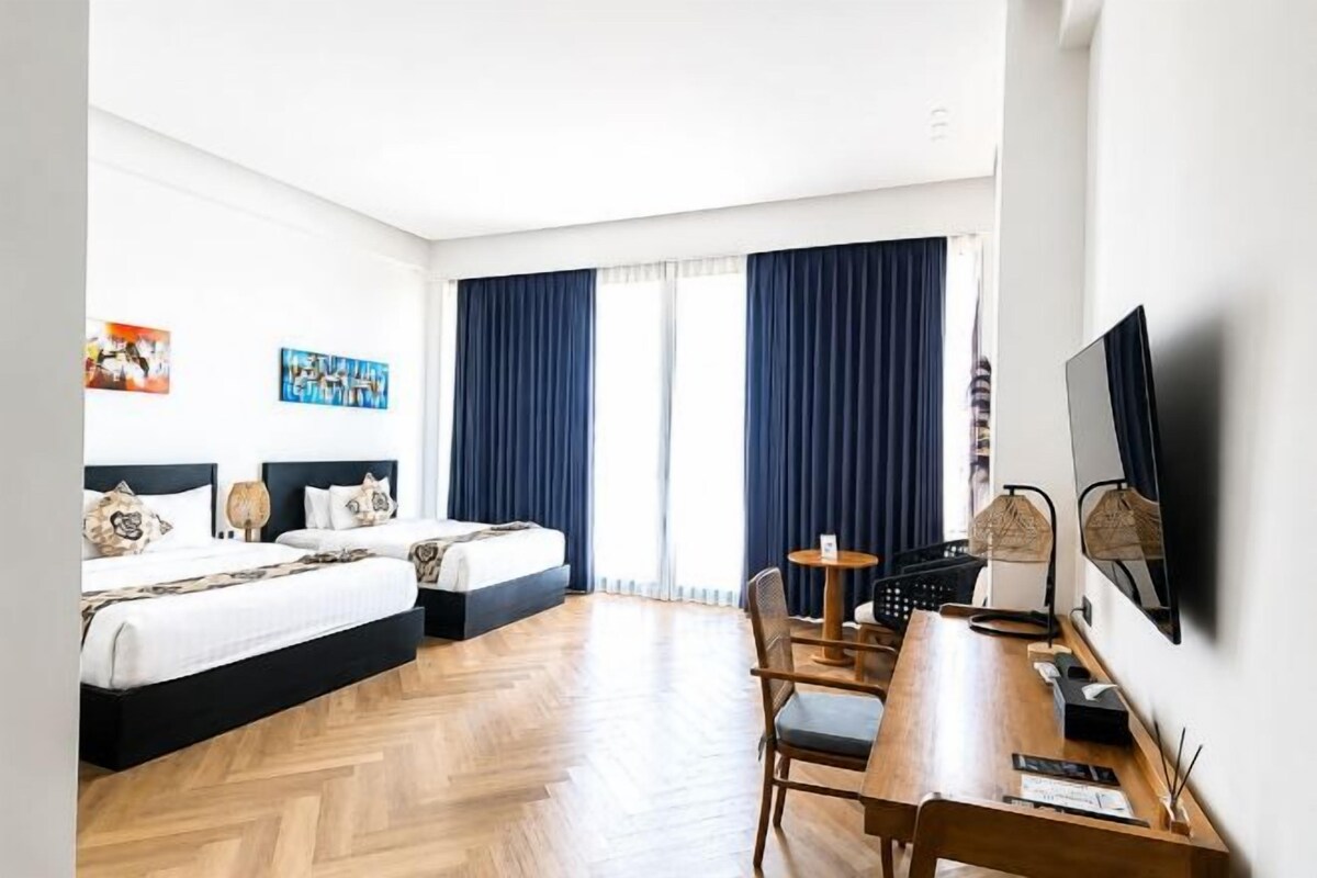 A hotel room with two beds, a desk, a chair, a TV, and a large window with curtains.