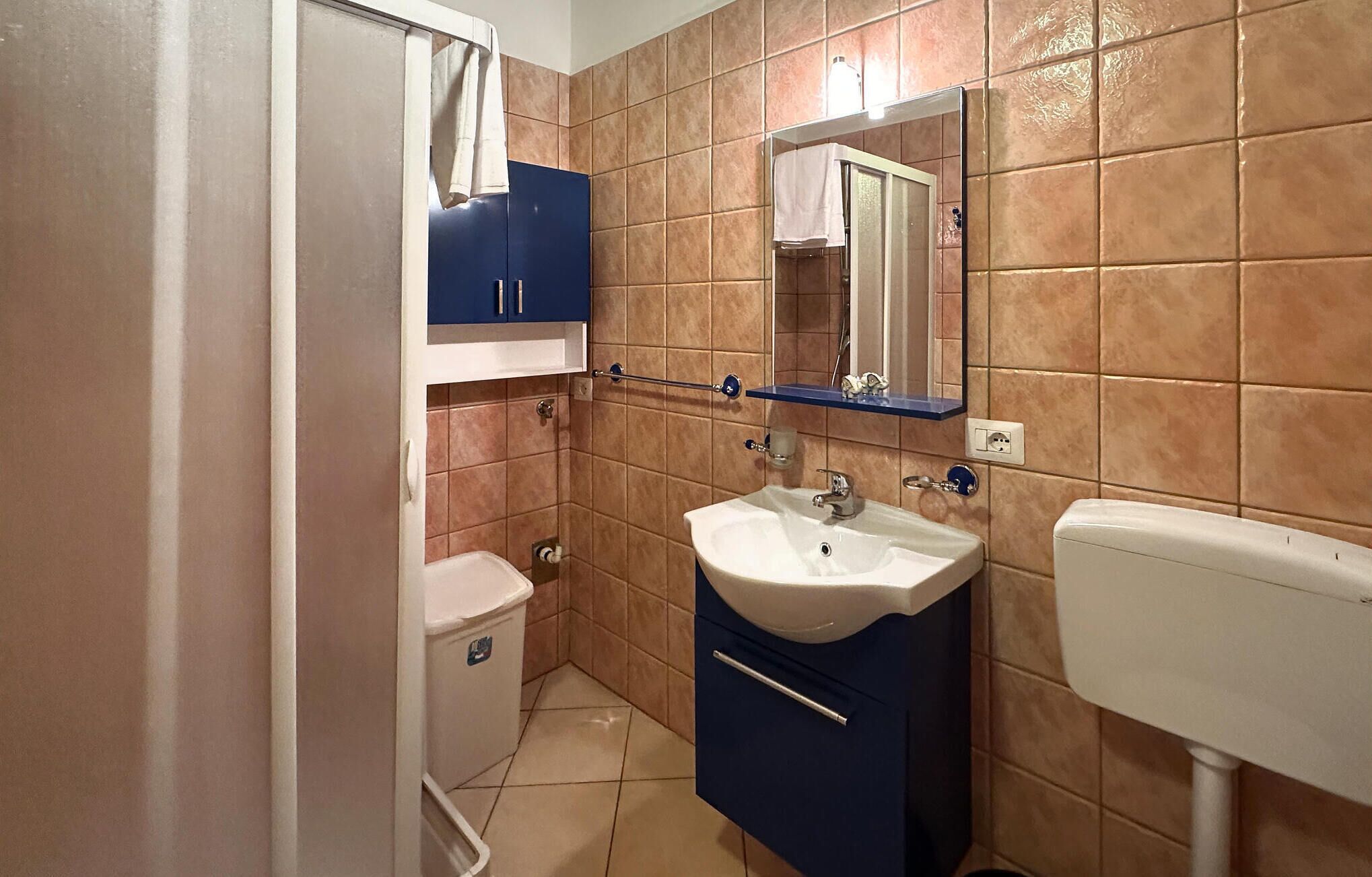 Une salle de bain avec des toilettes blanches, un lavabo surmonté d’un miroir et un meuble bleu.