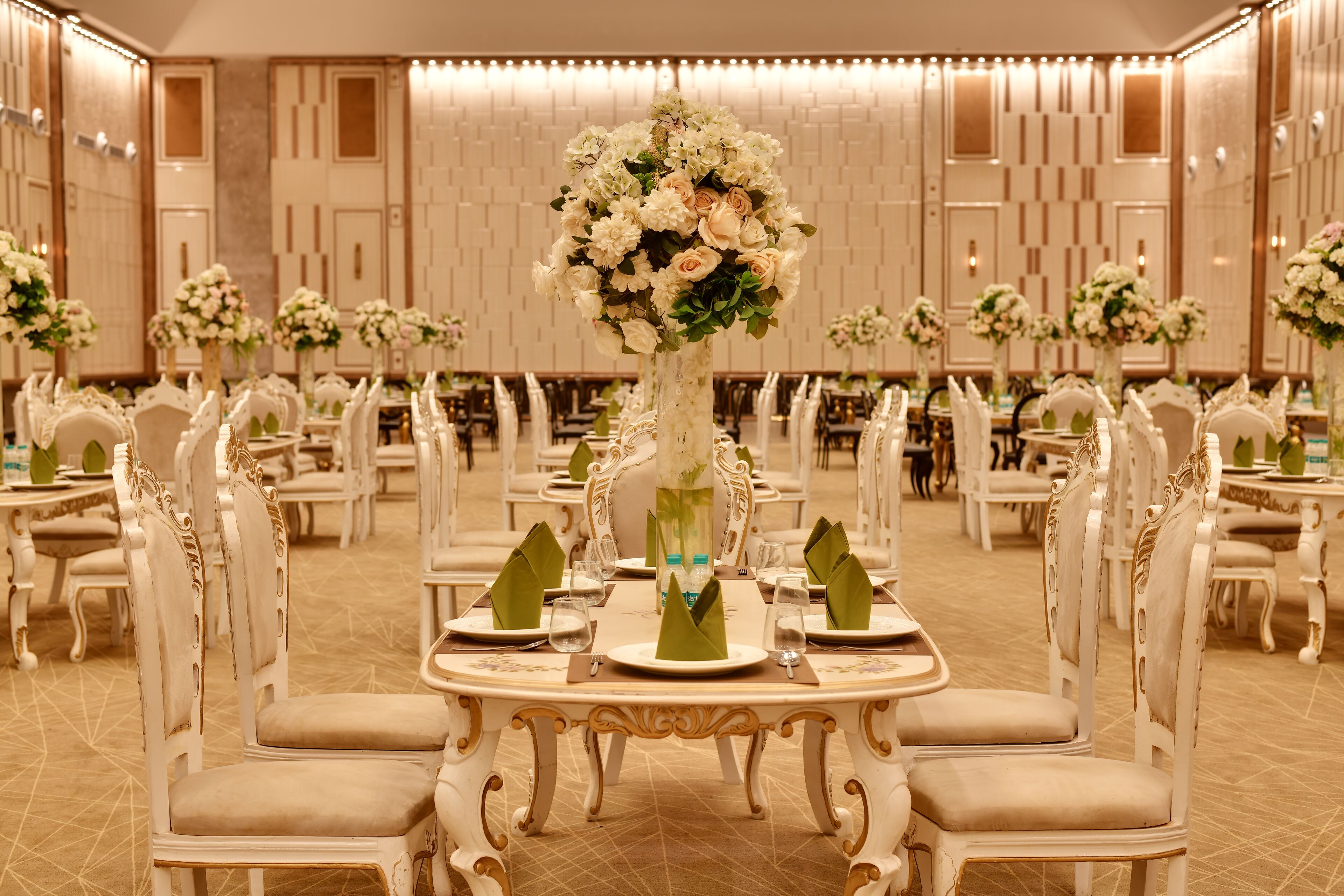 A banquet hall with ornate white chairs, round tables, and floral centerpieces.