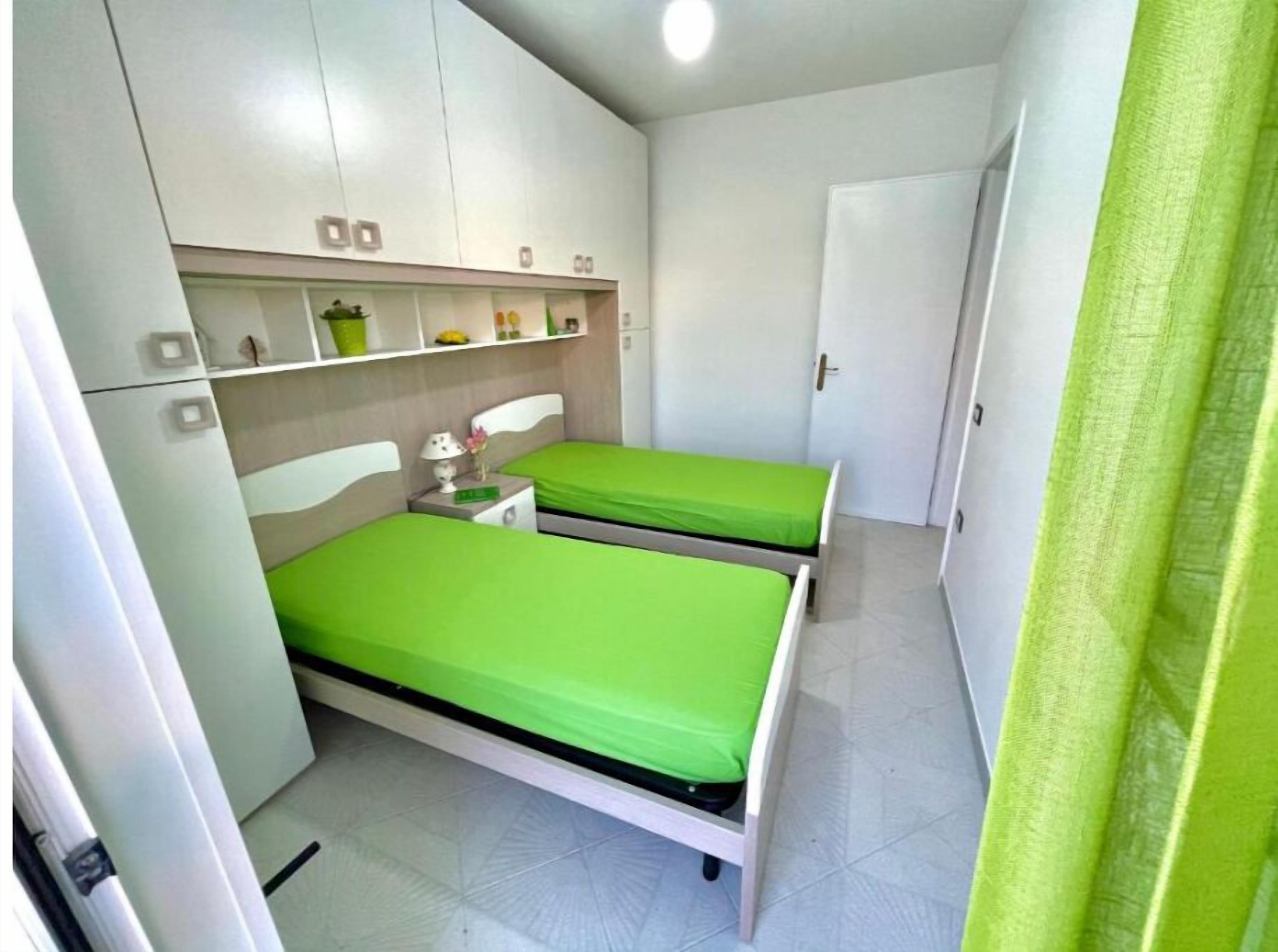A small, clean room with two single beds, a bedside table with a lamp, and a shelf with personal items.