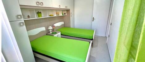 A small, clean room with two single beds, a bedside table with a lamp, and a shelf with personal items.