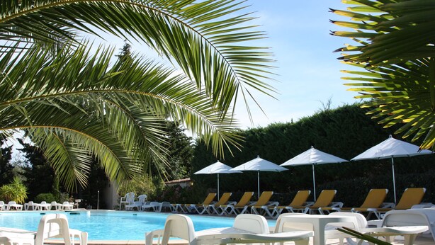 Un área de piscina con sillones de descanso y sombrillas, rodeada de palmeras y un seto.