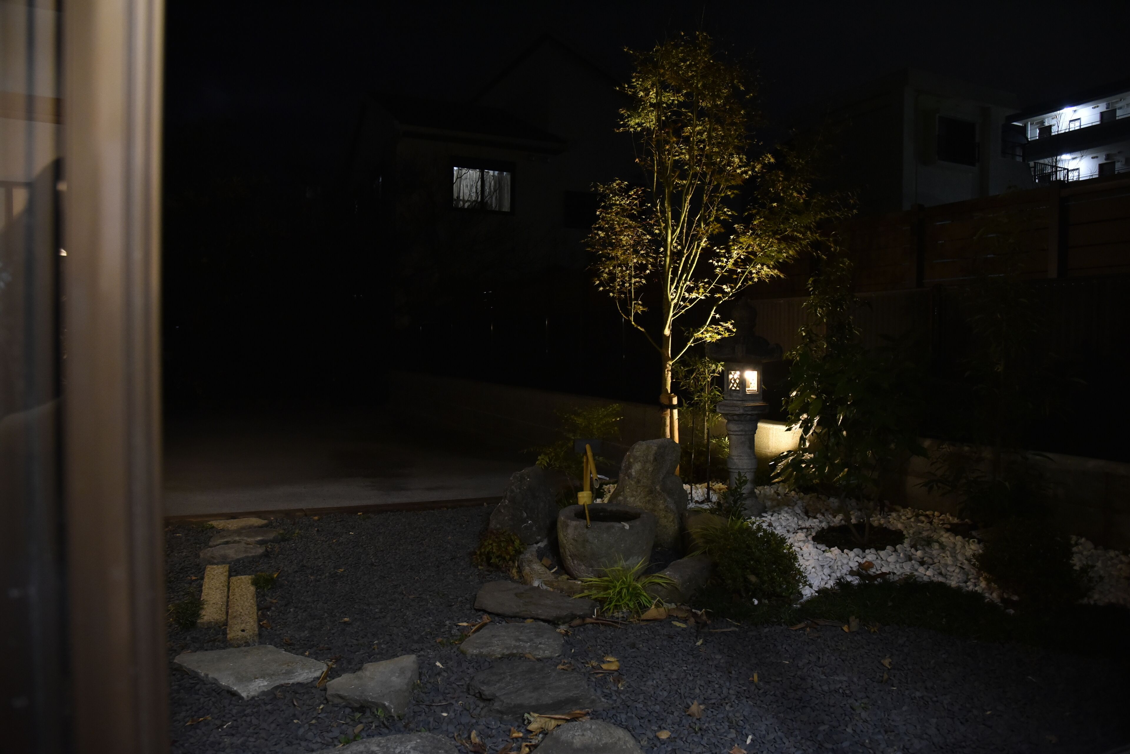 A nighttime view of a garden with a tree, rocks, and a building in the background.