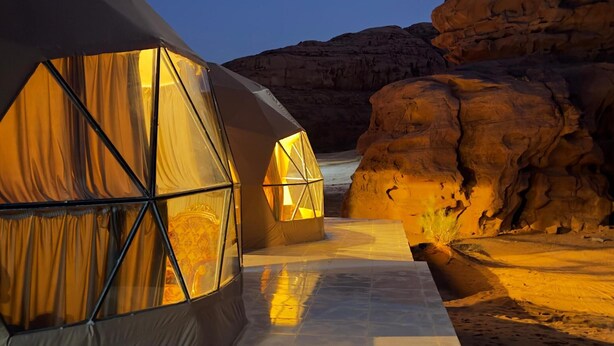 Acampamento no deserto com duas tendas geodésicas iluminadas por luzes internas quentes, com o relevo rochoso e o céu noturno ao fundo.
