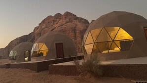 Três estruturas em forma de cúpula com grandes janelas, emolduradas por uma paisagem desértica e montanhas rochosas.