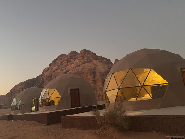 Three dome-shaped structures with large windows, set against a desert backdrop with rocky mountains.