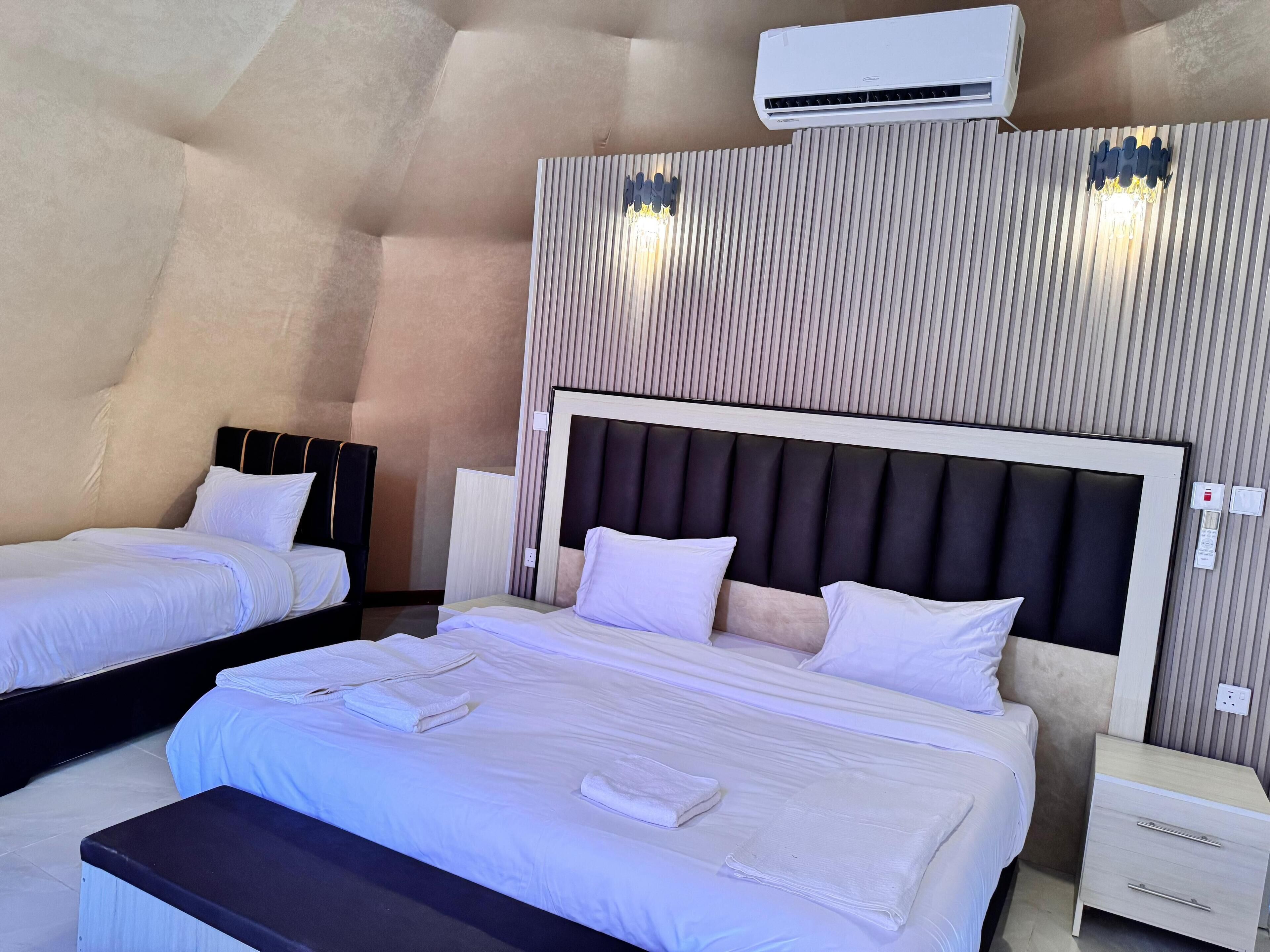 Habitación de hotel con dos camas, un banco, una mesita de noche, aire acondicionado y una lámpara fijada a la pared.