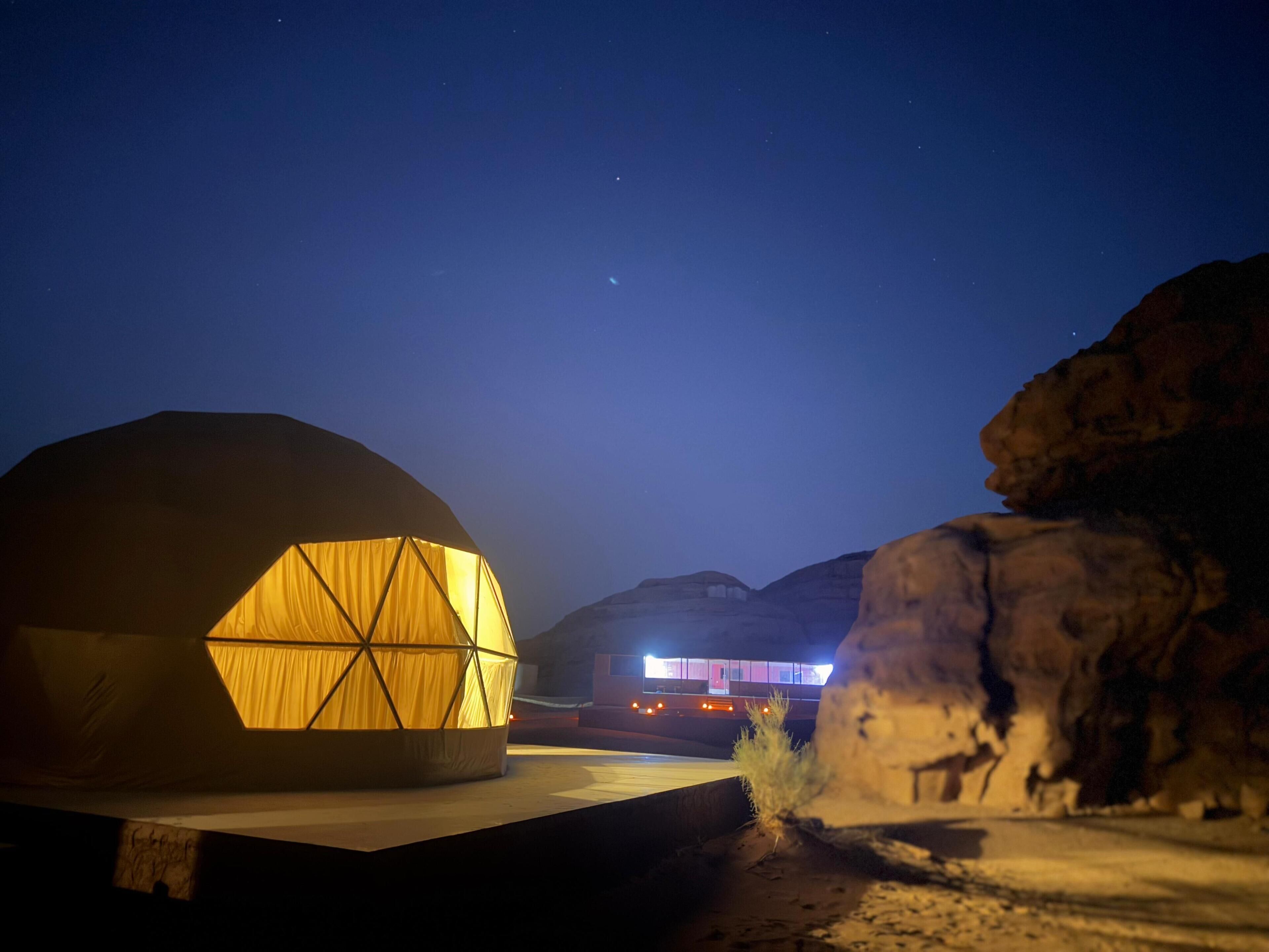 A geodesic dome structure with a warm interior light, set against a backdrop of mountains and a clear night sky.