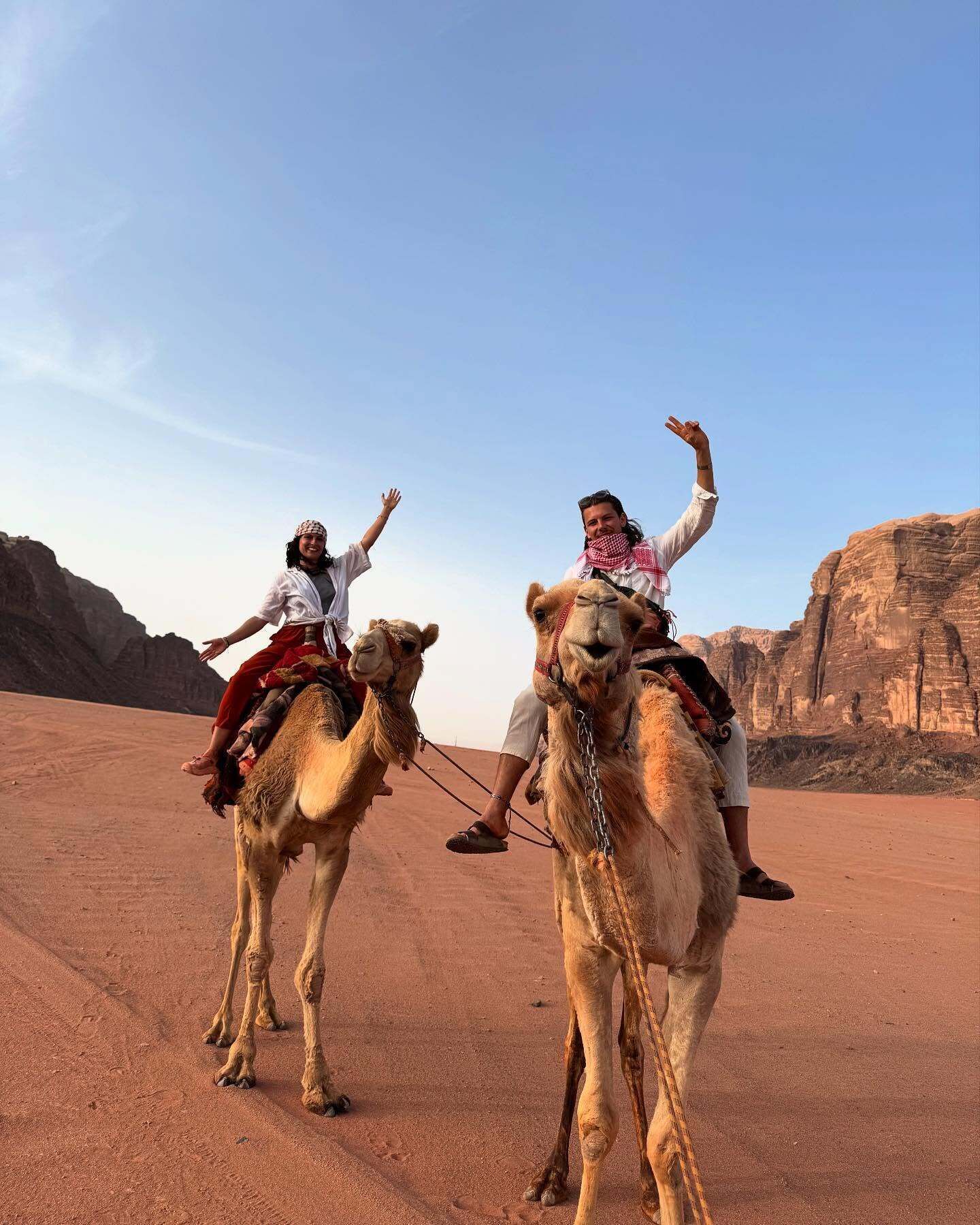 Two people riding camels in a desert landscape with rocky formations in the background.