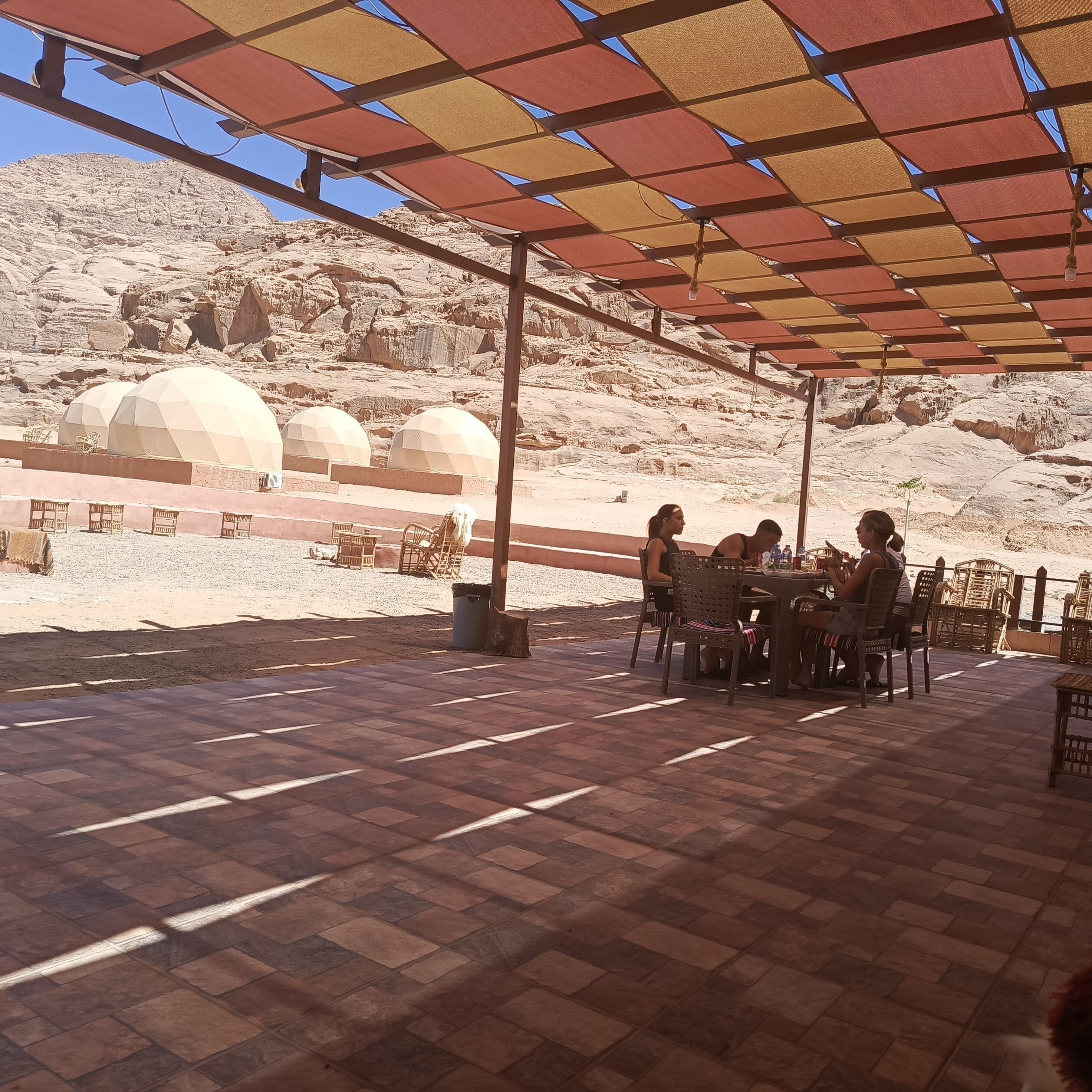 An outdoor seating area with tables and chairs, a covered walkway, and a view of a desert landscape with rock formations and domed structures.