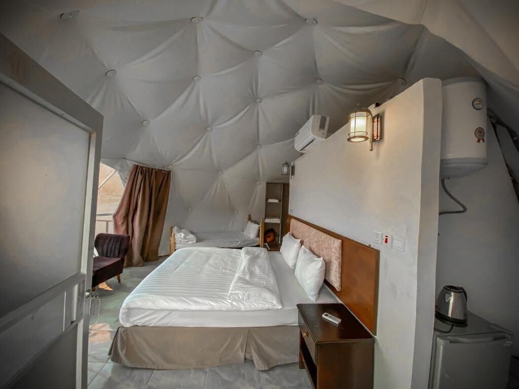Une pièce spacieuse avec un grand lit, une table de chevet en bois, un bureau et une chaise. On y trouve une fenêtre avec des rideaux, un climatiseur fixé au mur et une lampe fixée au mur.