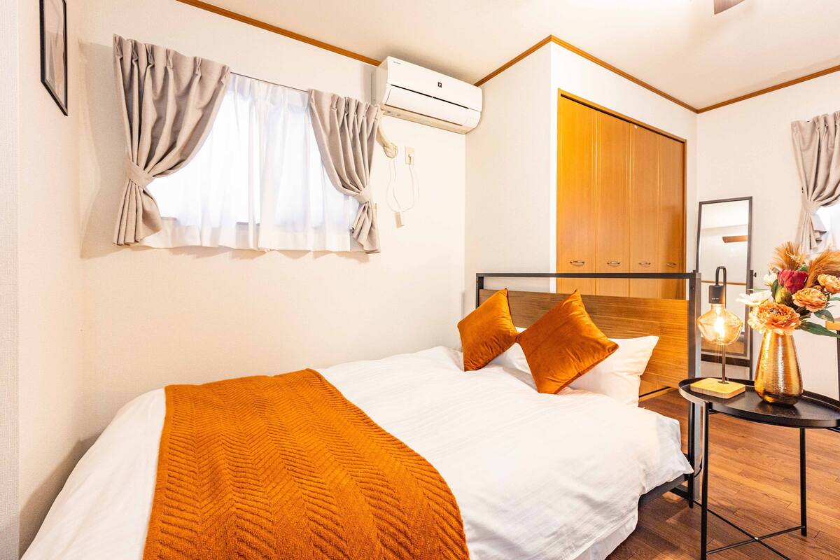 Ein Hotelzimmer mit einem Bett, Vorhängen, einer Klimaanlage, einem Holzschrank, einem Nachttisch mit Lampe und einer Vase mit Blumen.