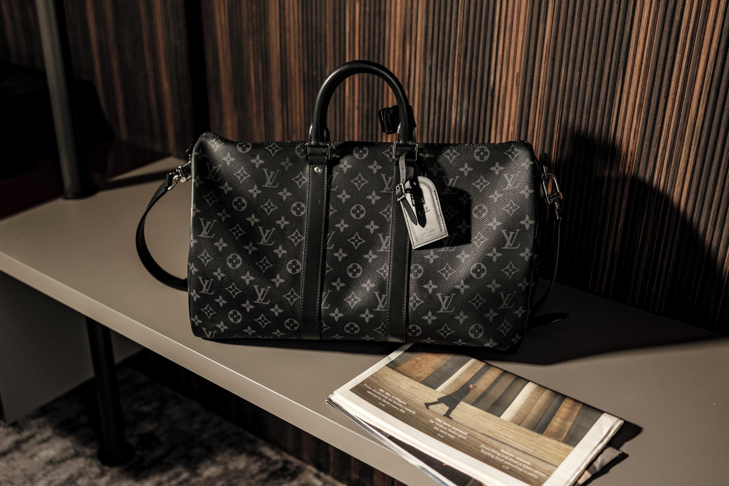 Musta Louis Vuitton -käsilaukku pöydällä, sen vieressä lehti.