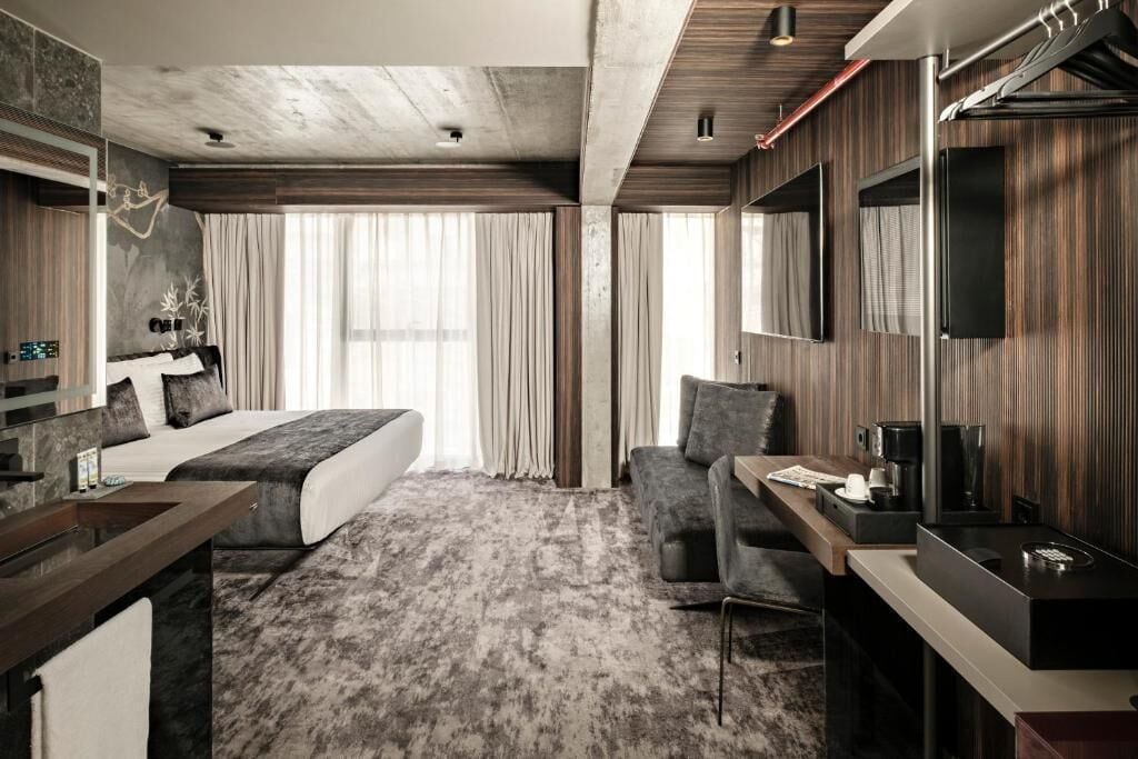 A modern hotel room with a bed, a desk, a chair, a TV, and a large window.
