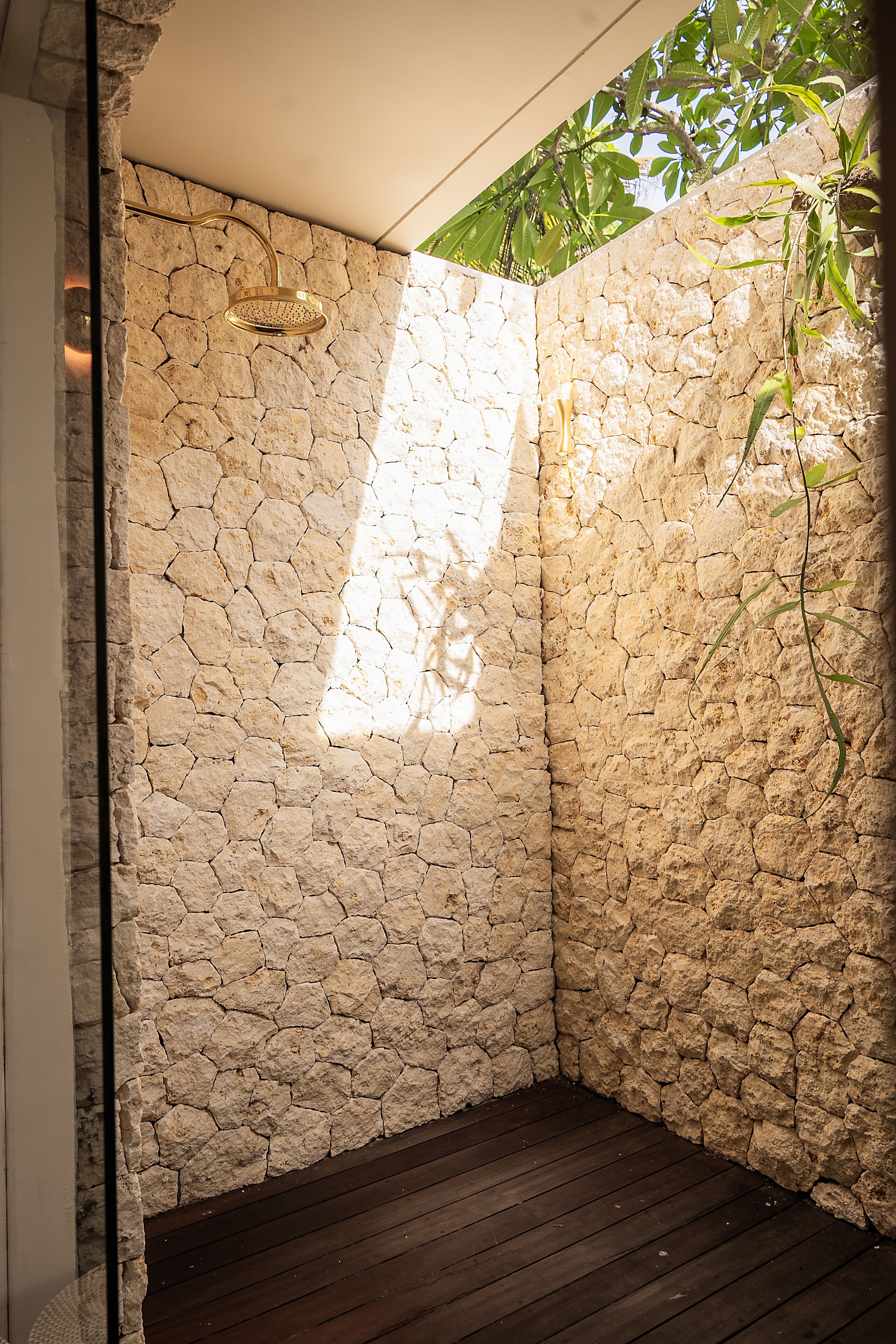 A stone-walled shower area with a wooden floor and a view of greenery outside.